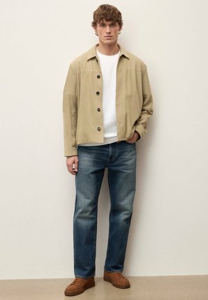 Młody mężczyzna w beżowej zapinanej na guziki kurtce, białej koszulce, niebieskich dżinsach i brązowych trampkach, stojący na tle jednolitej jasnej ściany.