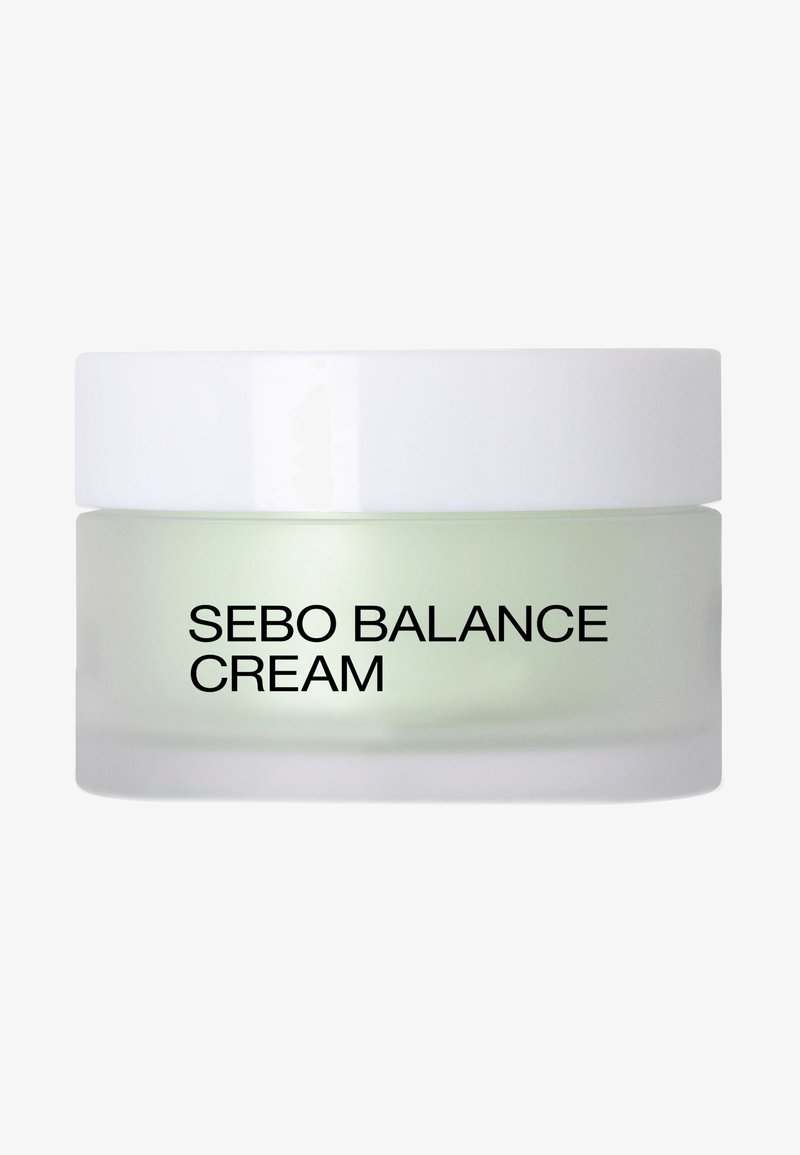Crème Sebo Balance dans un pot en verre givré avec un couvercle blanc ; crème verdâtre visible à l'intérieur ; texte noir en gros sur l'étiquette avant.