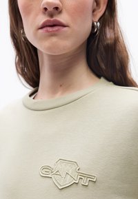 Κοντινό πλάνο μιας γυναίκας που φοράει ανοιχτό μπεζ φούτερ με κεντητό λογότυπο GANT στο στήθος και ασημένια κρίκους σκουλαρίκια, με λευκό φόντο.