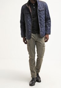 Quiltet marineblå jakke med brune detaljer, sort poloshirt, olivengrønne cargobukser med sidelommer og sorte snørestøvler.