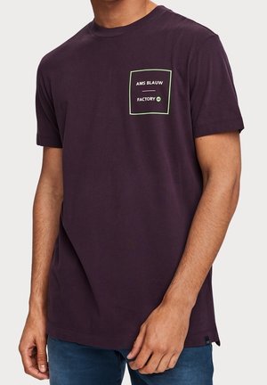 Mężczyzna ubrany w ciemnofioletowy t-shirt z krótkim rękawem z nadrukiem na kieszeni na piersi obrysowanym na zielono z napisem "AMS BLAUW FACTORY AB".