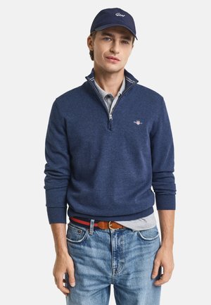 Marineblauwe ritspullover met geribde mouwen en kraag, gecombineerd met een lichtgrijze polo, gedragen met lichtblauwe jeans en een bruine riem.