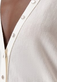 Vaaleanbeige neulottu neuletakki, V-pääntie-muotoilu, etupuolella hopeiset painonapit, jossa on teksturoitu kangas, jossa on kevyt kiilto.