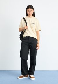 Beige t-shirt med "DICKIES" trykt i sort, sorte bukser og brune sneakers med hvide såler. Modellen holder en lille sort crossbody taske.