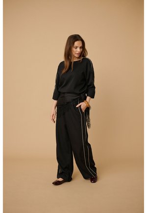 Kobieta stojąca na beżowym tle, ubrana w luźne czarne spodnie z białymi paskami po bokach, czarny top i brązową bransoletkę, patrzy w dół.