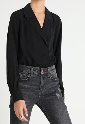 Persoon draagt een zwarte blouse met lange mouwen en kraag, ingestopt in hoog getailleerde verweerde zwarte jeans met een kleine beschadigde plek op het bovenbeen.