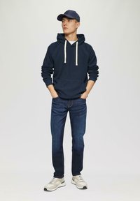 Sudadera azul marino con cordones crema, bolsillo frontal; jeans de mezclilla oscura; zapatillas grises con acentos verdes y blancos; gorra de béisbol.