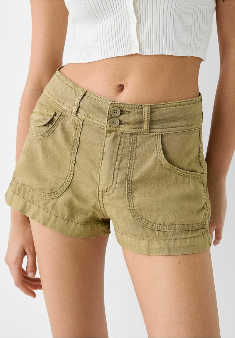 Shorts en khaki avec une texture douce, taille haute, poches avant et fermeture à double bouton. Présente des ourlets arrondis et des coutures contrastées.