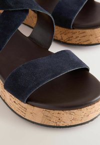 Mornarško modri suede platform sandali s srednjico iz plute, ki imajo široke naramnice in gladek, temno rjav podplat. Teksturosana pluta dodaja kontrast.