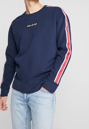 Sweatshirt bleu marine à col rond, doté d'un patch logo central et d'accents rayés rouge et blanc le long des manches.