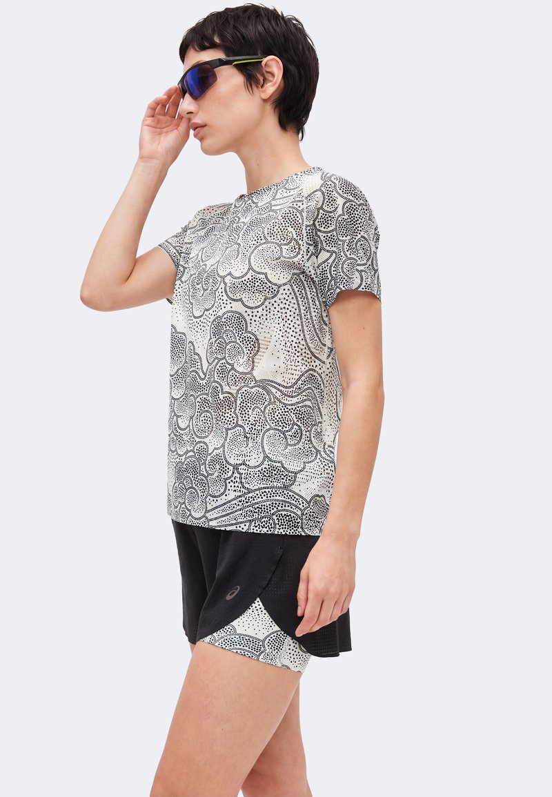 Frau passt blau getönte Sonnenbrille an, trägt ein gemustertes weiß-schwarzes Kurzarmhemd und schwarze Shorts mit passender gemusterter Innenlage.