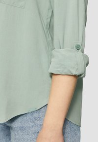 Lichtgroene blouse van een zachte, gestructureerde stof. Voorzien van opgerolde mouwen die zijn vastgezet met knopen en een gebogen zoom. Denimstof zichtbaar eronder.