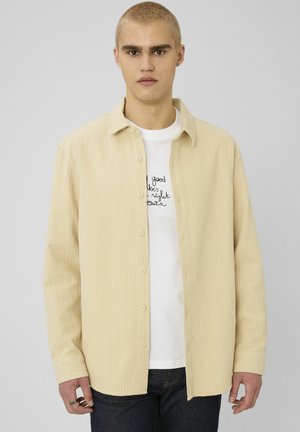 Jeune homme aux cheveux blonds courts portant une chemise beige boutonnée sur un t-shirt graphique blanc et un jean foncé, debout devant un fond uni.