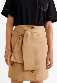 Falda envolvente color caqui con detalle de cinturón anudado, acento de botón de madera y suave tela de algodón, combinada con una camiseta negra de manga corta.