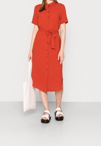 Abito rosso con bottoni e maniche corte, con cintura in vita e taglio dritto. Indossato con sandali bianchi e mentre tiene una borsa tote di un rosa chiaro.