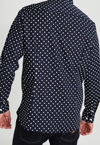 Marineblauwe broek met witte stippen, lange mouwen, een button-down kraag en een rechte snit. De stof lijkt glad en lichtgewicht.