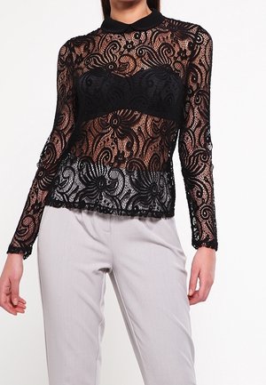 Frau trägt ein schwarzes langärmeliges Spitzenshirt mit floralem Muster und einem einfarbigen schwarzen Kragen, kombiniert mit hellgrauen maßgeschneiderten Hosen.