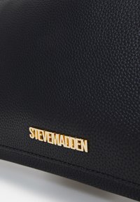 Borsa nera testurizzata con logo dorato "STEVE MADDEN" e dettagli di cucitura liscia. Presenta un design strutturato e materiali resistenti.