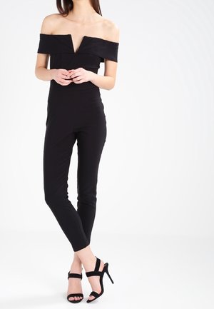 Nainen pukeutuneena mustaan, olkapäät paljastavaan, vartalonmyötäiseen haalariin, jonka lahkeet ovat nilkkapituiset, ja mustiin korkokenkiin, seisomassa ristityin jaloin.