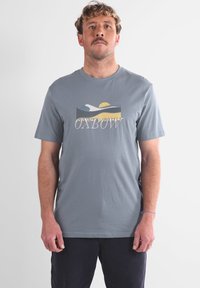 Homme avec des cheveux frisés courts et une moustache, portant un T-shirt gris avec un design de vagues jaune, blanc et bleu et le texte "OXBOW", debout.