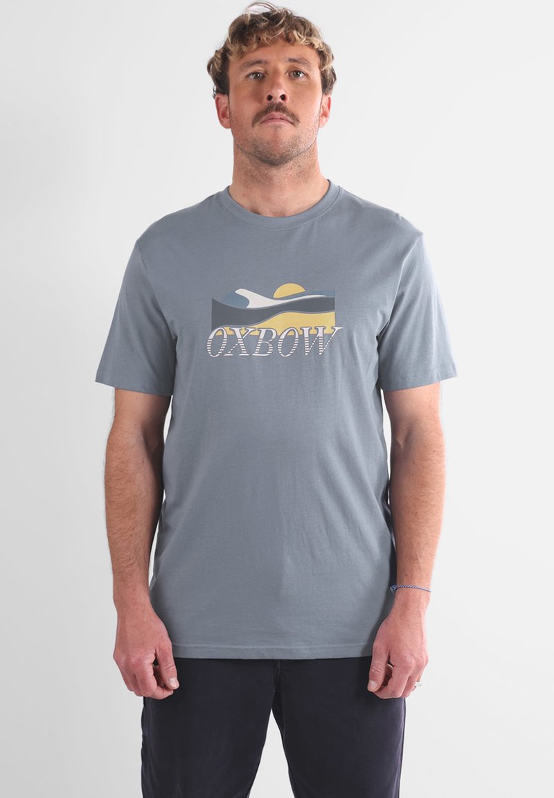 Homme avec des cheveux frisés courts et une moustache, portant un T-shirt gris avec un design de vagues jaune, blanc et bleu et le texte "OXBOW", debout.