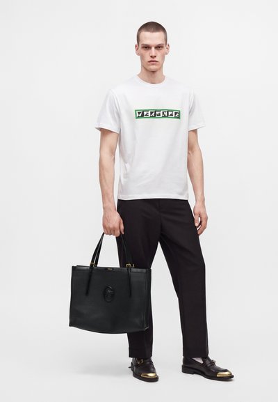 Homme portant un t-shirt blanc Versace, pantalon noir, mocassins noirs avec embouts dorés, tenant un grand sac en cuir noir avec l'emblème de Méduse.