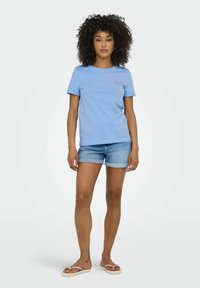 Camiseta de manga corta azul claro, con cuello redondo, que presenta un sutil emblema en forma de corazón. Combinada con pantalones cortos de mezclilla enrollados y chanclas. Textura suave.