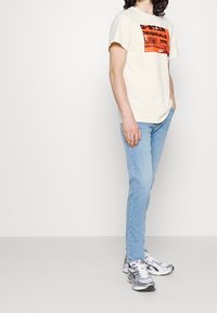 Camiseta beige con gráfico negro y naranja, jeans ajustados azul claro y zapatillas deportivas grises con acentos negros y amarillos.