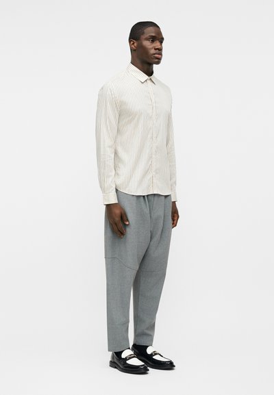 Άνδρας που στέκεται φορώντας εκρού πουκάμισο με ρίγες και κουμπιά, γκρι παντελόνι φαρδιάς γραμμής, μαύρα και λευκά loafers σε απλό φωτεινό φόντο.