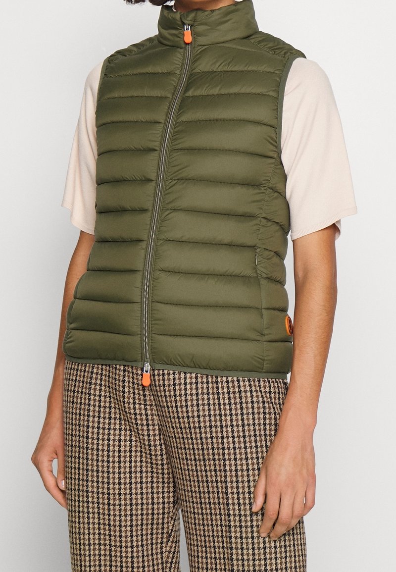 Zielona oliwkowa kamizelka puchowa z wysokim kołnierzem, poziomym przeszyciem i pomarańczowym uchwytem zamka. Nosi ją nad jasnym beżowym topem i wzorzystymi brązowymi spodniami.
