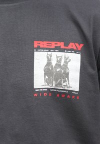 Γκρι βαμβακερό μπλουζάκι με γραφικό σχέδιο τριών σκυλιών, με έντονο κόκκινο κείμενο "REPLAY" και "WIDE AWAKE" να προβάλλεται κάτω.