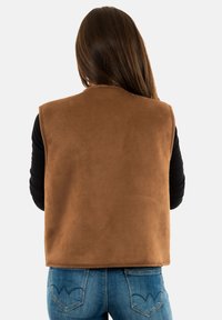 Gilet in camoscio marrone con colletto alto, design senza maniche e trama liscia, indossato sopra una maglietta a maniche lunghe nera e abbinato a jeans blu.