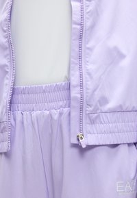 Svetlofialová bunda a nohavice s elastickým pásom v rovnakej farbe s logom EA7 Emporio Armani na nohaviciach pri zipsu.
