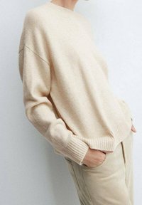 Pull en maille beige avec un col rond, des poignets côtelés et un ourlet côtelé. Texture douce et coupe décontractée. Associé à un pantalon beige clair.
