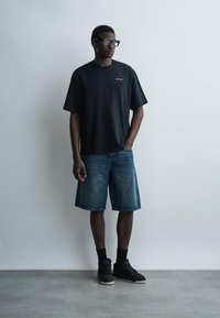 Mees kannab üle suurte musta t-särgi, põlvpikkuseid teksapükse, musti sokke, tossusid ja prille, seisab ühetaolise heleda seina ees.