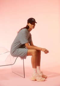 Gestreiftes, kurzärmliges Kleid in Schwarz und Weiß, getragen mit einer schwarzen Kap. Dazu pinkfarbene Turnschuhe und weiße Socken, sitzt auf einem transparenten Stuhl vor einem rosa Hintergrund.