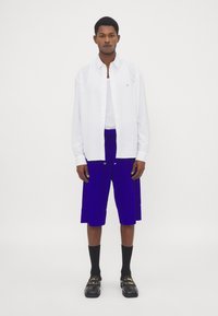 Witte overhemd met knopen, blauwe shorts met een trekkoord in de taille, zwarte sokken en schoenen met gouden details. Eenvoudig en eigentijds ontwerp.