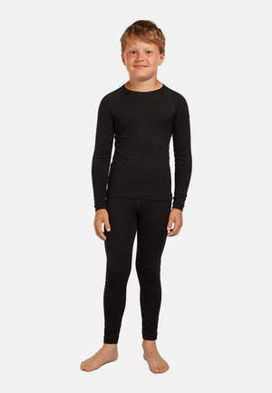 Ragazzo in piedi a piedi nudi che indossa una maglia nera a maniche lunghe e leggings neri abbinati su uno sfondo bianco uniforme.