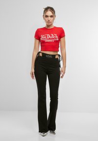 Červené zkrácené tričko s bílým logem "Von Dutch Originals", doplněné černými rozšířenými kalhotami. Kalhoty mají boční tkaničky a pás s logem.
