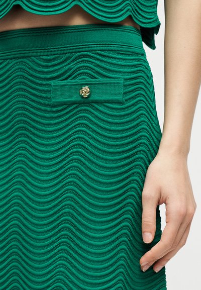 Gros plan sur une jupe verte texturée avec un motif ondulé, une petite poche à rabat avec un bouton doré, et une main posée à côté.