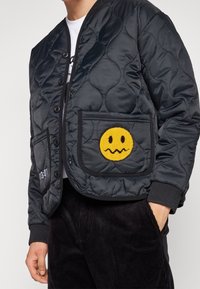 Veste matelassée noire avec fermeture à boutons et poignets côtelés, arborant un grand motif de smiley jaune sur la poche droite.
