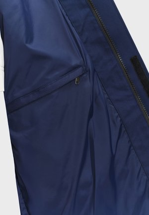 Intérieur d'une veste bleu marine montrant une poche zippée, un curseur de fermeture éclair noir et une bande auto-agrippante noire sur le bord de la doublure.