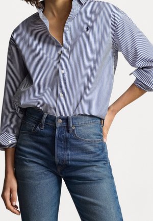 Femme portant une chemise à rayures verticales bleues et blanches à boutons, manches retroussées, rentrée dans un jean taille haute bleu, main sur la hanche.