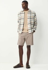Lässiges Outfit mit einem karierten Hemd in Creme-, Braun- und Blautönen, einem beigen T-Shirt, hellbraunen Shorts und dunkle braunen Slippers.