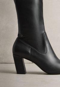 Svart läderstövel med fyrkantig klack, slät yta, minimal sömnad och elegant design, med avsmalnande tå och hög skaft.