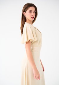 Beiges Kleid mit kurzen, flatternden Ärmeln. Glattes, leichtes Material, das sanft fällt. Tailliertes Oberteil, das zu einem fließenden Rock führt.