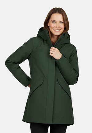 Vrouw draagt een donkergroene jas met capuchon en klepzakken, lacht en sluit de ritssluiting aan de voorkant.