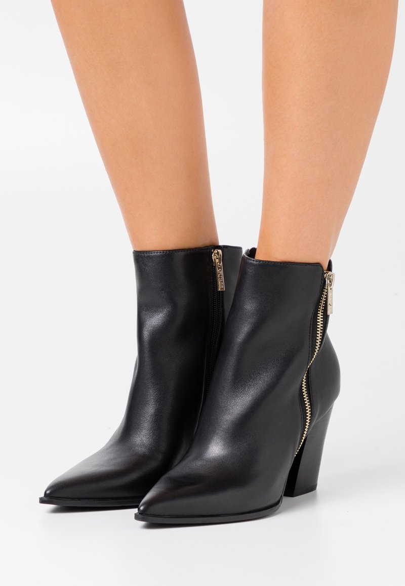 Bottines en cuir noir avec fermetures éclair dorées sur les côtés et hauts talons blocs portées sur jambes nues sur fond blanc.