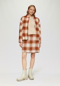 Braun- und cremefarbener Karojacke, passendem Rock und strukturiertem beigen Pullover, kombiniert mit cremefarbenen Stiefeletten mit einer klobigen Sohle.