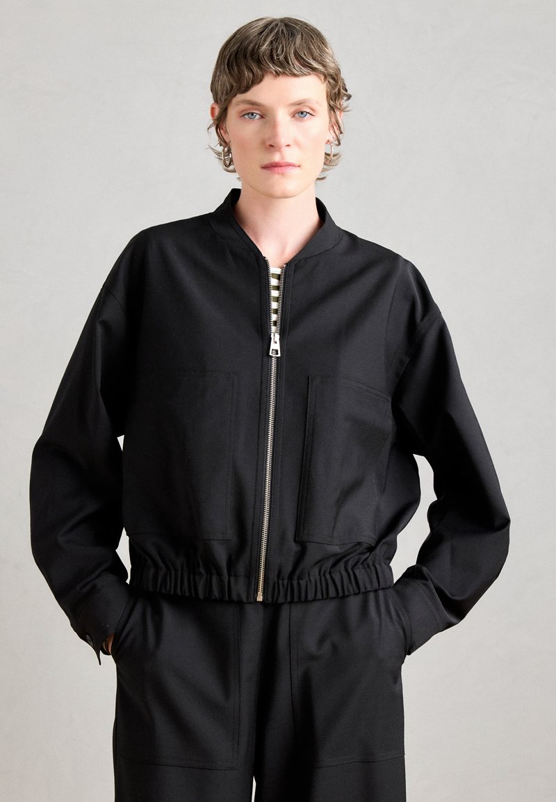 Veste noire zippée avec col haut et taille élastique. Dispose de deux poches avant et d'une texture de tissu lisse. Doublure intérieure rayée visible.
